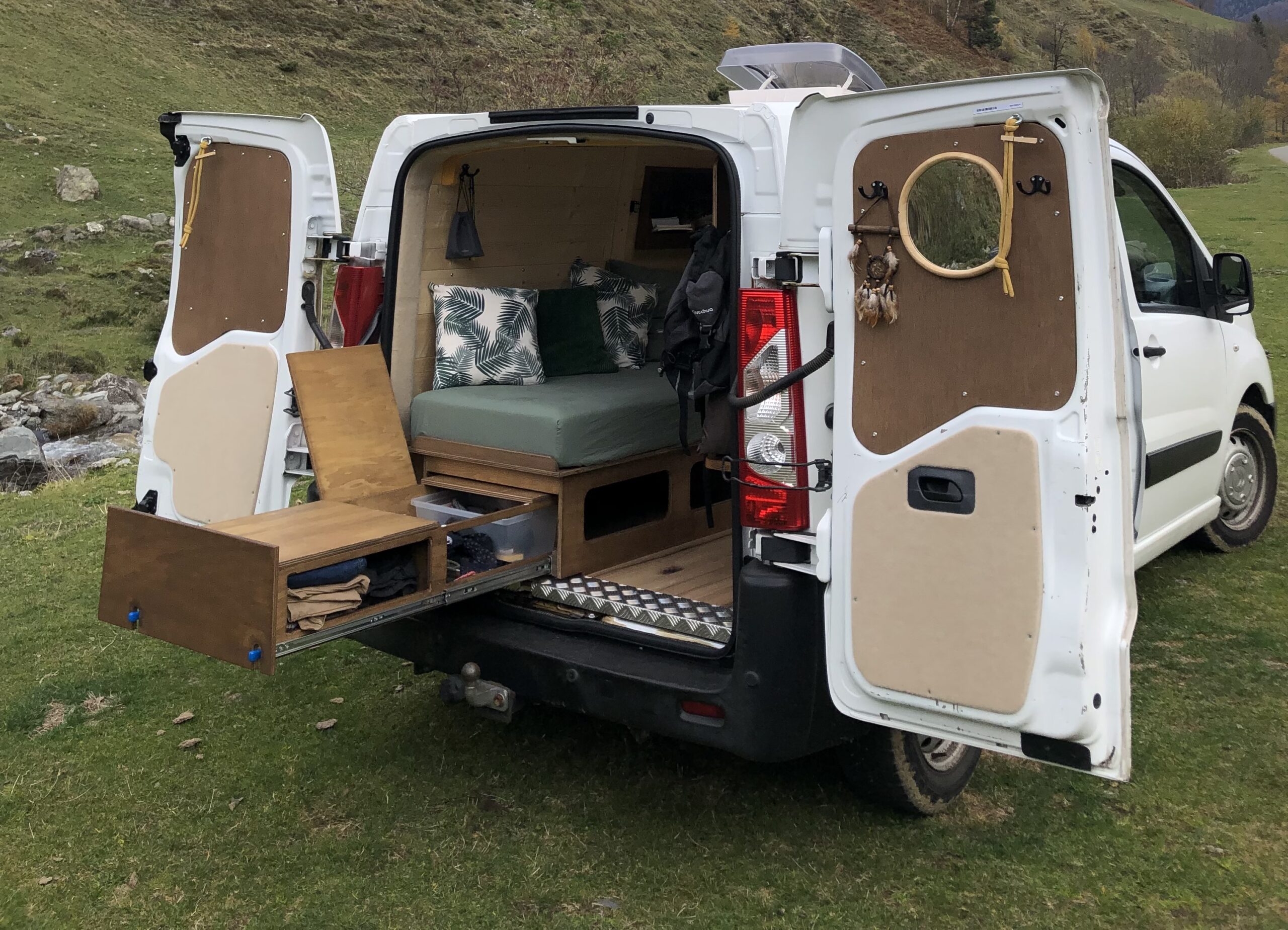 Aménagement intérieur d’un van aménagé avec du bois, disposant d'un lit peigne, d'un coin cuisine, rangements et espace cosy pour la vie en van